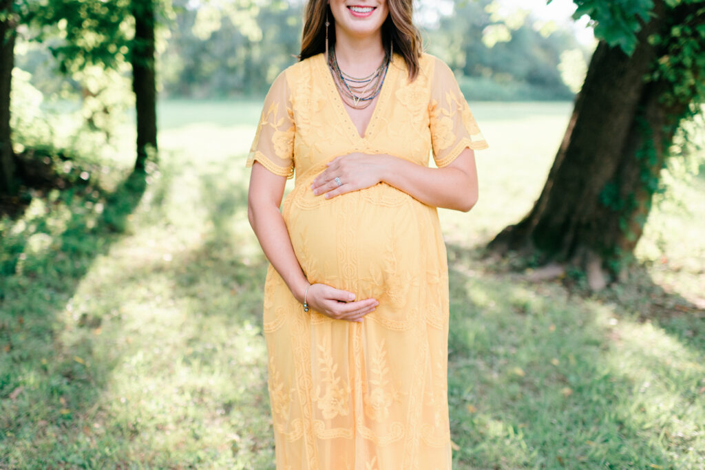 Bentonville Maternity photography session detail shot of a pregnant woman’s hands resting on her belly, framed by a natural backdrop of trees and field.