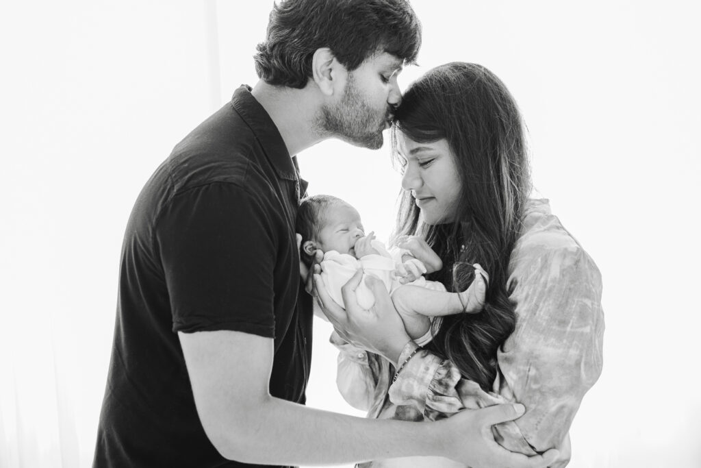 A heartfelt black-and-white photograph of new parents cradling their newborn baby. The father kisses the mother gently on the forehead while she gazes lovingly at the baby, creating an intimate and tender family moment during their Bentonville newborn session.