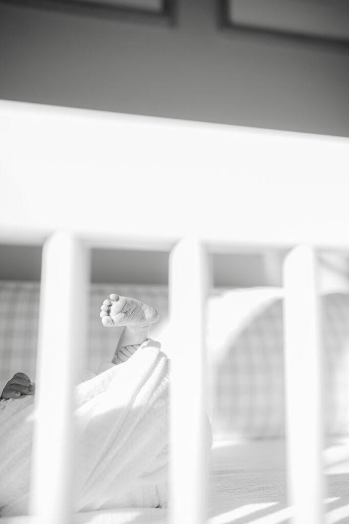 A tiny foot peeks through the crib slats. Newborn baby swaddled in a blanket, lying in crib in softly lit nursery during an in-home newborn photography session in Bentonville.