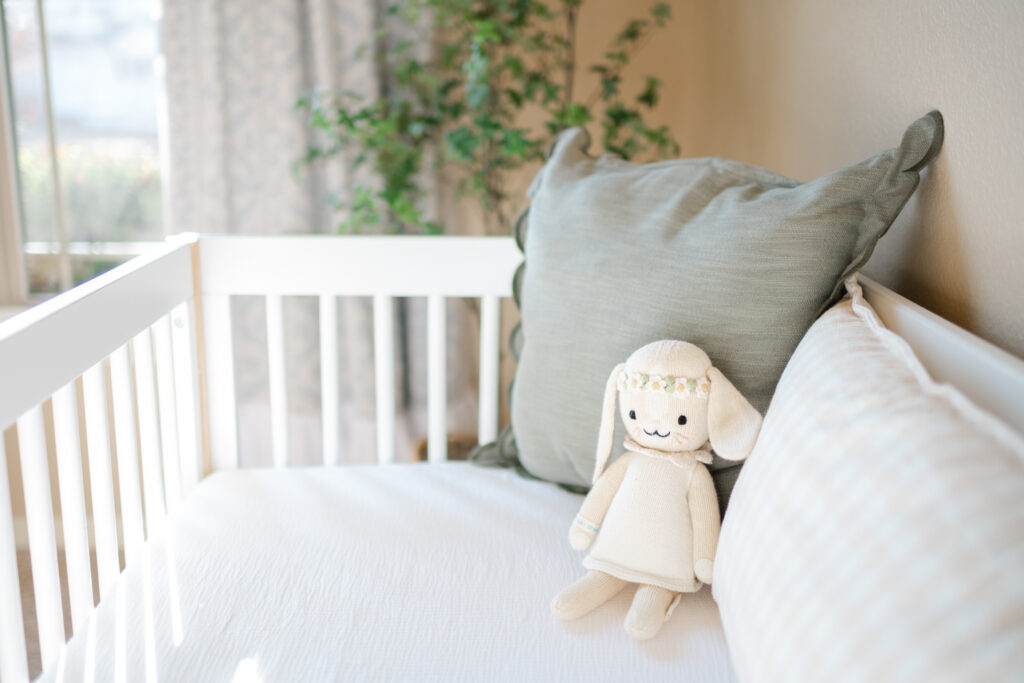 A white crib adorned with a soft, knit stuffed bunny wearing a flower crown, set against a serene nursery backdrop with a green pillow and natural light streaming through the window, captured during an in-home newborn photography session in Bentonville.
