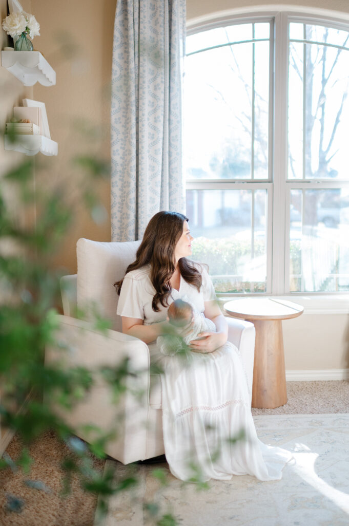 Mother in a white dress cradling her newborn baby in a softly lit nursery with a large window, highlighting the natural light and peaceful atmosphere of an in-home newborn photography session in Bentonville