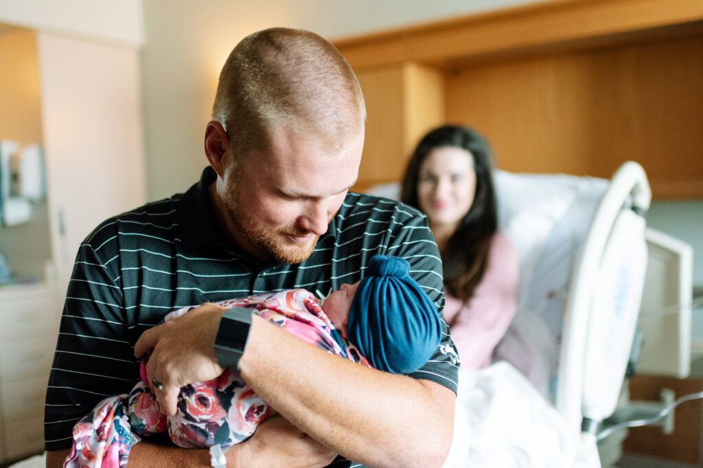 Mom looks on as dad holds newborn baby in hospital room. NWA Maternity Photographer.