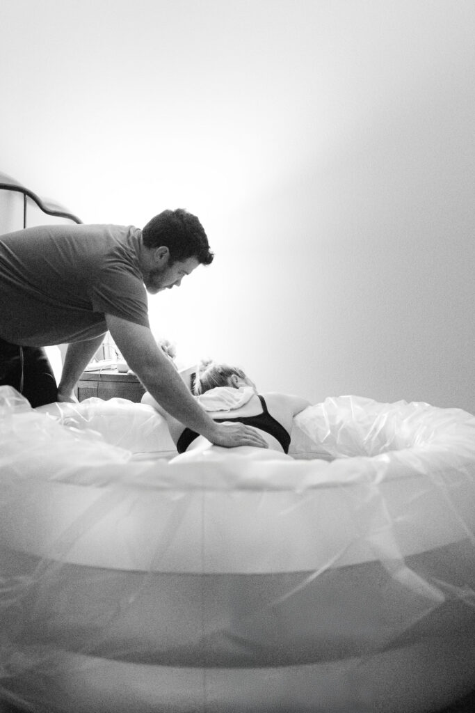 Woman labors in waterbirth tub while her husband places a hand on her back in support. 