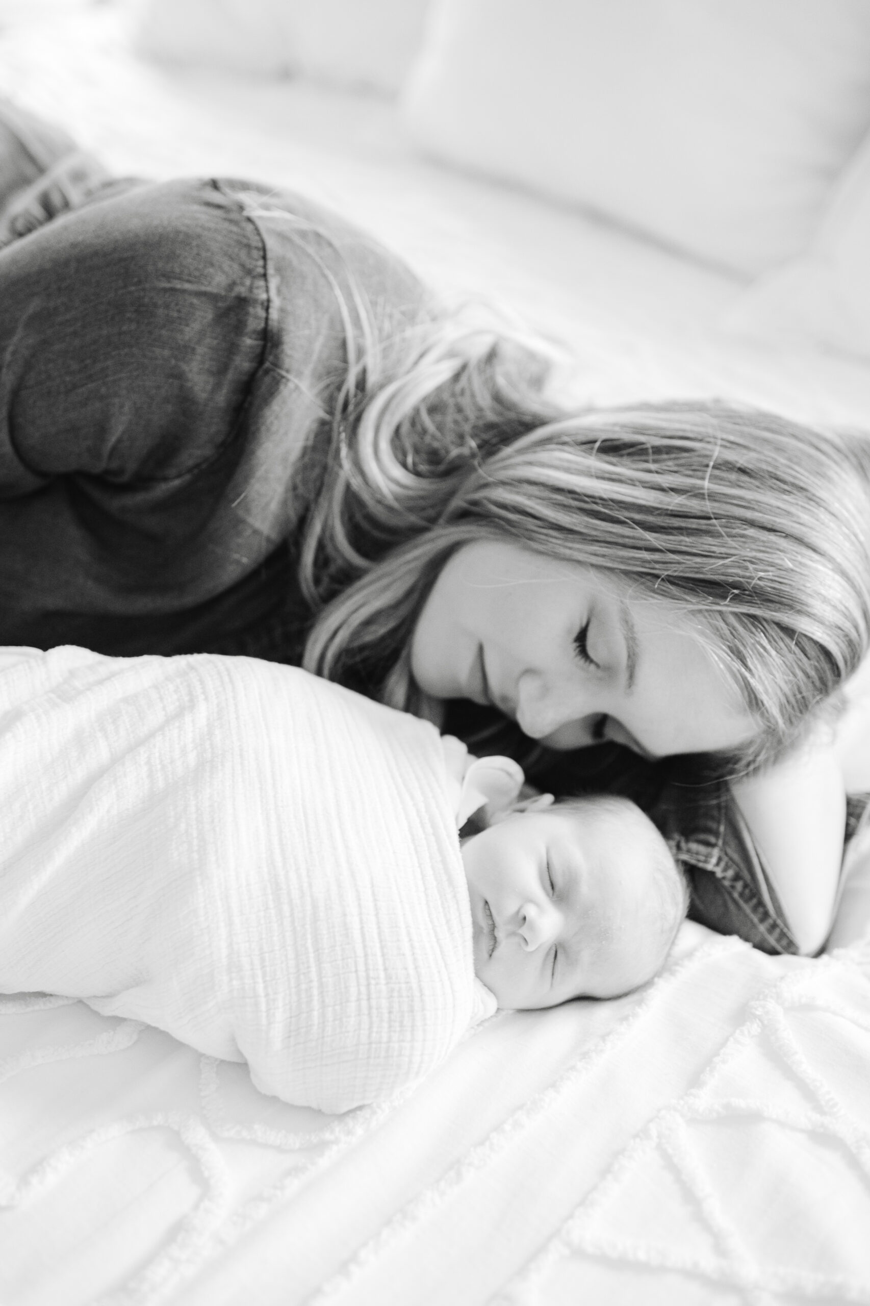 Black and white photograph by nwa photographer Leslie Nice. Photo of a mother lying on a bed, lovingly gazing at her swaddled newborn baby who is peacefully asleep