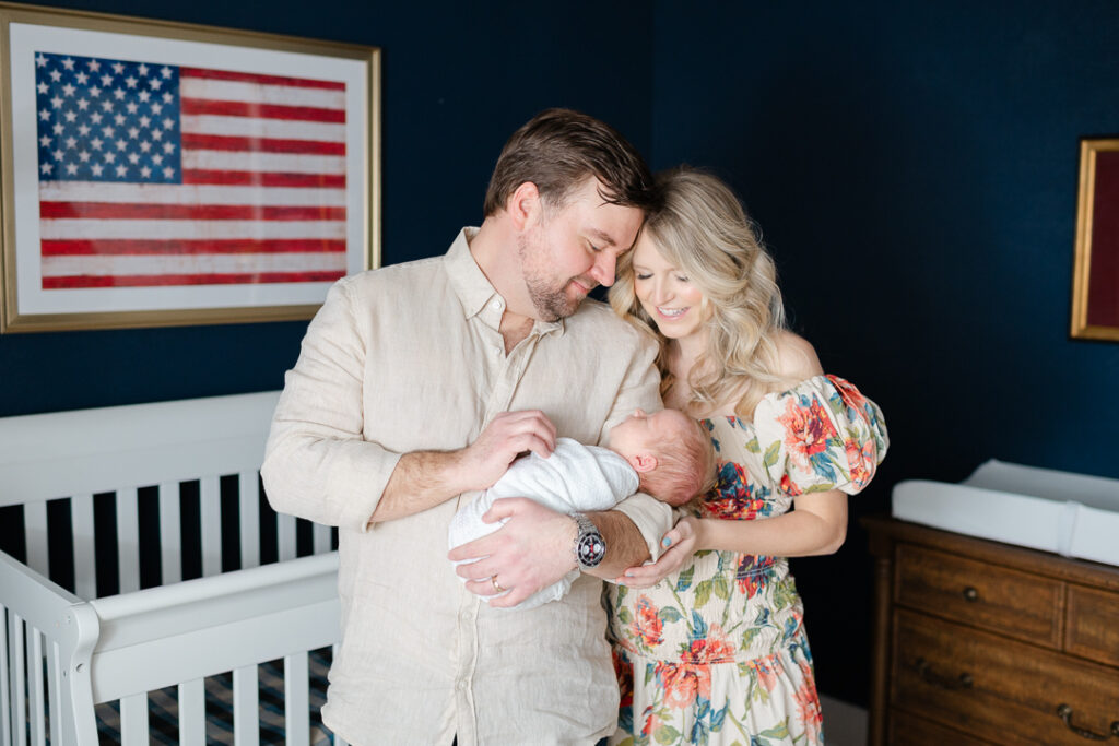 A mother and father stand together in front of a white crib, cradling their newborn baby. A framed American flag hangs on the wall above the crib, adding a sentimental touch to the nursery. Captured by one of the talented NWA photographers, this image beautifully preserves the emotions of new parenthood and the love shared in this special moment.