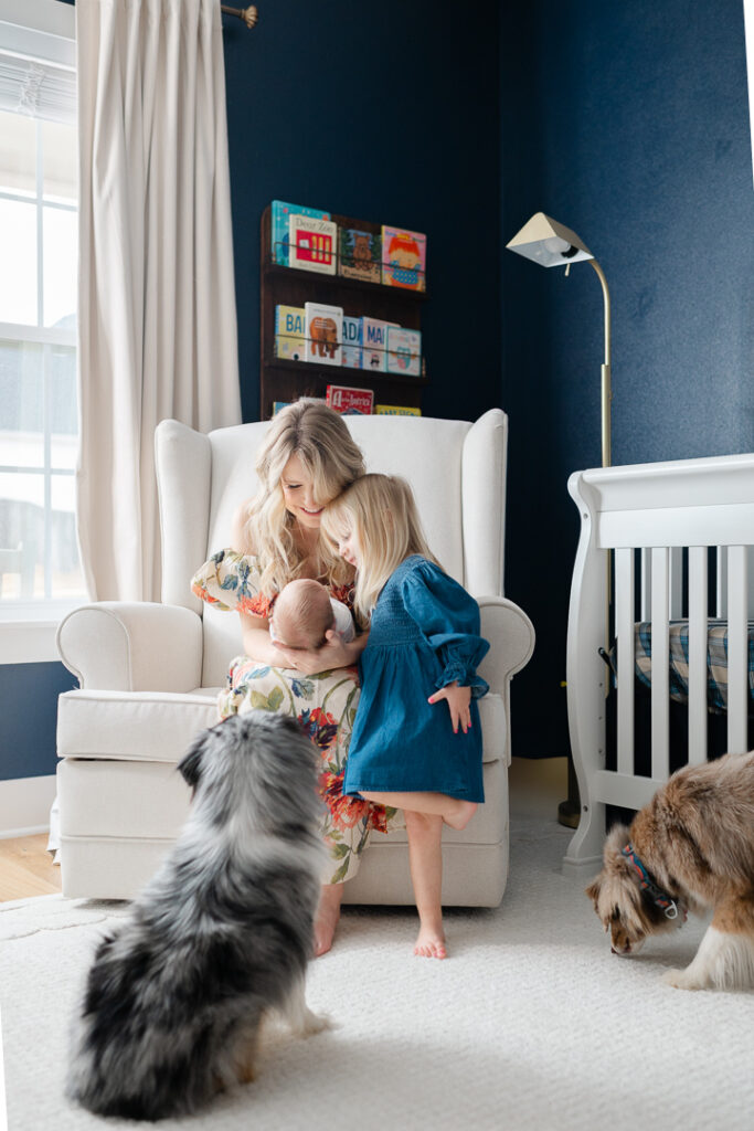 A mother sits in a rocking chair near a bright window, holding her newborn while her toddler daughter looks on with curiosity and love. Two family dogs rest in the foreground, completing this warm and intimate scene. This lifestyle newborn session, photographed by one of the skilled NWA photographers, highlights the beauty of everyday family moments filled with love, connection, and natural light.