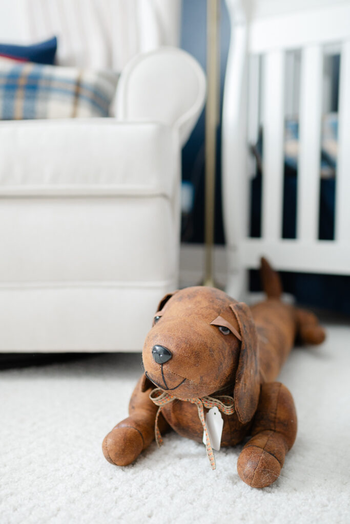 A stuffed animal wiener dog on the floor, a white crib and ivory rocking chair are blurred in the background. Nursery details captured by a talented NWA newborn photographer. 