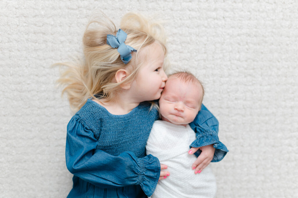 A sweet moment featuring a newborn baby snuggled in his big sisters arms, lying on a softly textured surface. The baby’s serene expression and carefully styled setup evoke a feeling of warmth and comfort. This lifestyle newborn session, photographed by one of the skilled NWA photographers, highlights the beauty of everyday family moments filled with love, connection, and natural light.