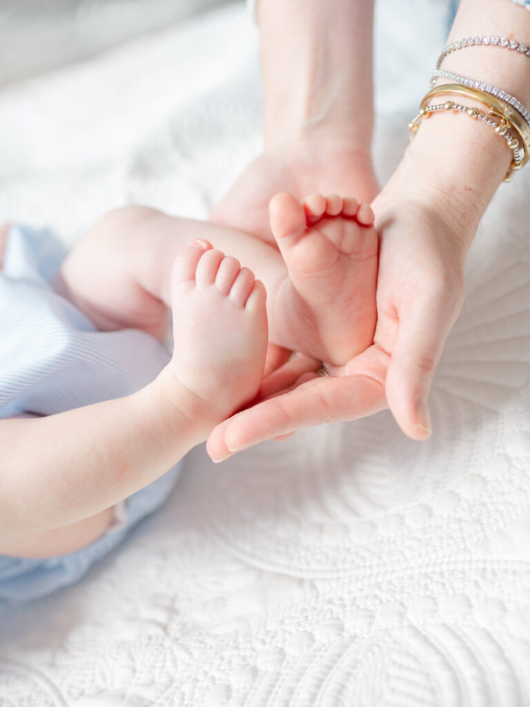 Mother's hands holding newborn feet near a window with soft light — example of connection-focused newborn photography at home.