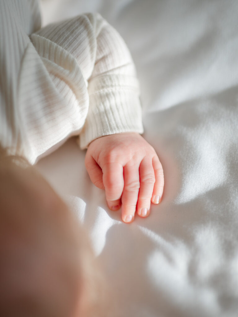 Close up of newborn hand, background of soft bed with neutral linens and a clean background for a minimal, timeless look.